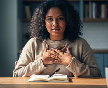 A diverse person seated at a table holding an open journal, symbolizing sharing lived experiences for mental health support.